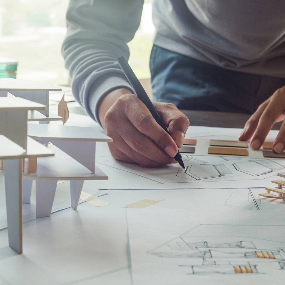 Architect draws on blueprints next to miniature building models, selecting material samples. ZUK-Architecture. Architect draws on blueprints next to miniature building models, selecting material samples. ZUK-Architecture.