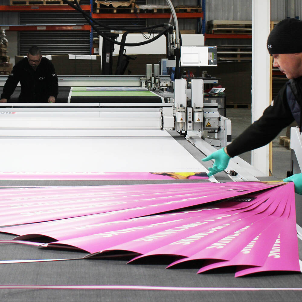 A Zünd cutter precisely trims pink printed materials, with a worker in teal gloves guiding the process. A Zünd cutter precisely trims pink printed materials, with a worker in teal gloves guiding the process.