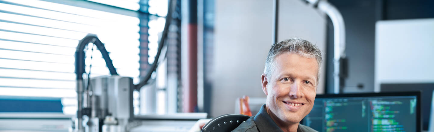 Ein lächelnder Mitarbeiter von Zünd Systemtechnik sitzt im Büro vor einem Computer und einer Schneidemaschine. Ein lächelnder Mitarbeiter von Zünd Systemtechnik sitzt im Büro vor einem Computer und einer Schneidemaschine.