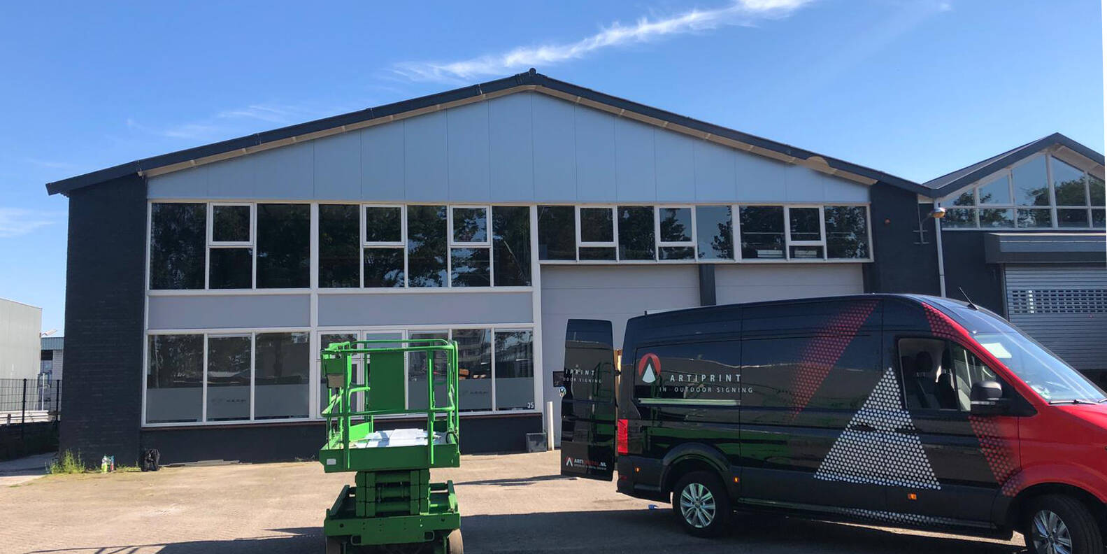 Een groene hoogwerker staat voor een gebouw met grote ramen en een bestelwagen met het opschrift 'ARTIPRINT'.