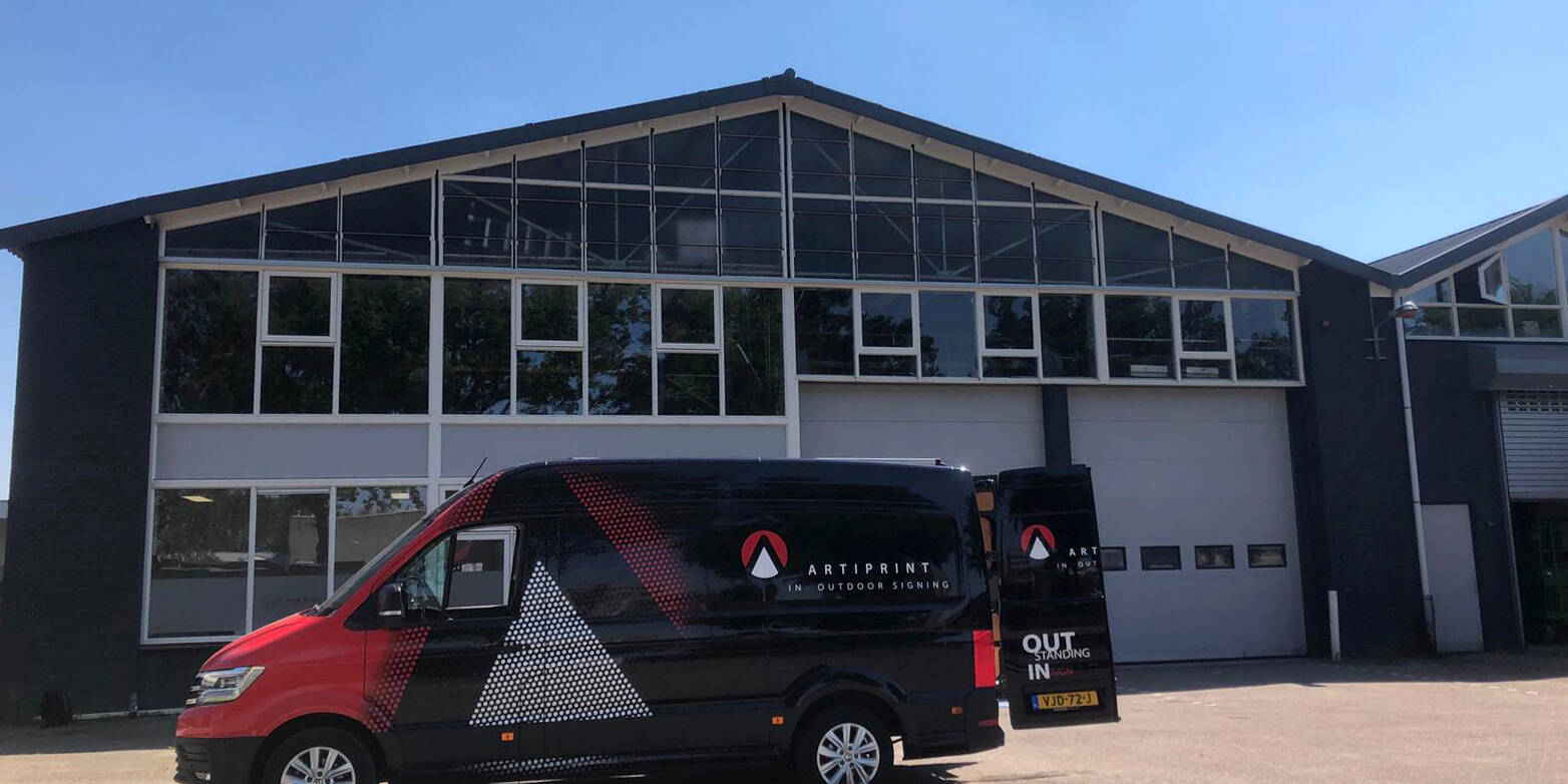 Bedrijfsbus van Artiprint voor buitensignalisatie geparkeerd voor een modern gebouw met grote ramen.