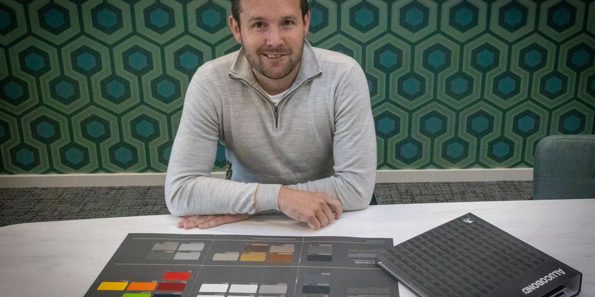 Man in grijs gebreid vest zit achter een tafel met een Alucobond kleurenpalet en monstertafel.