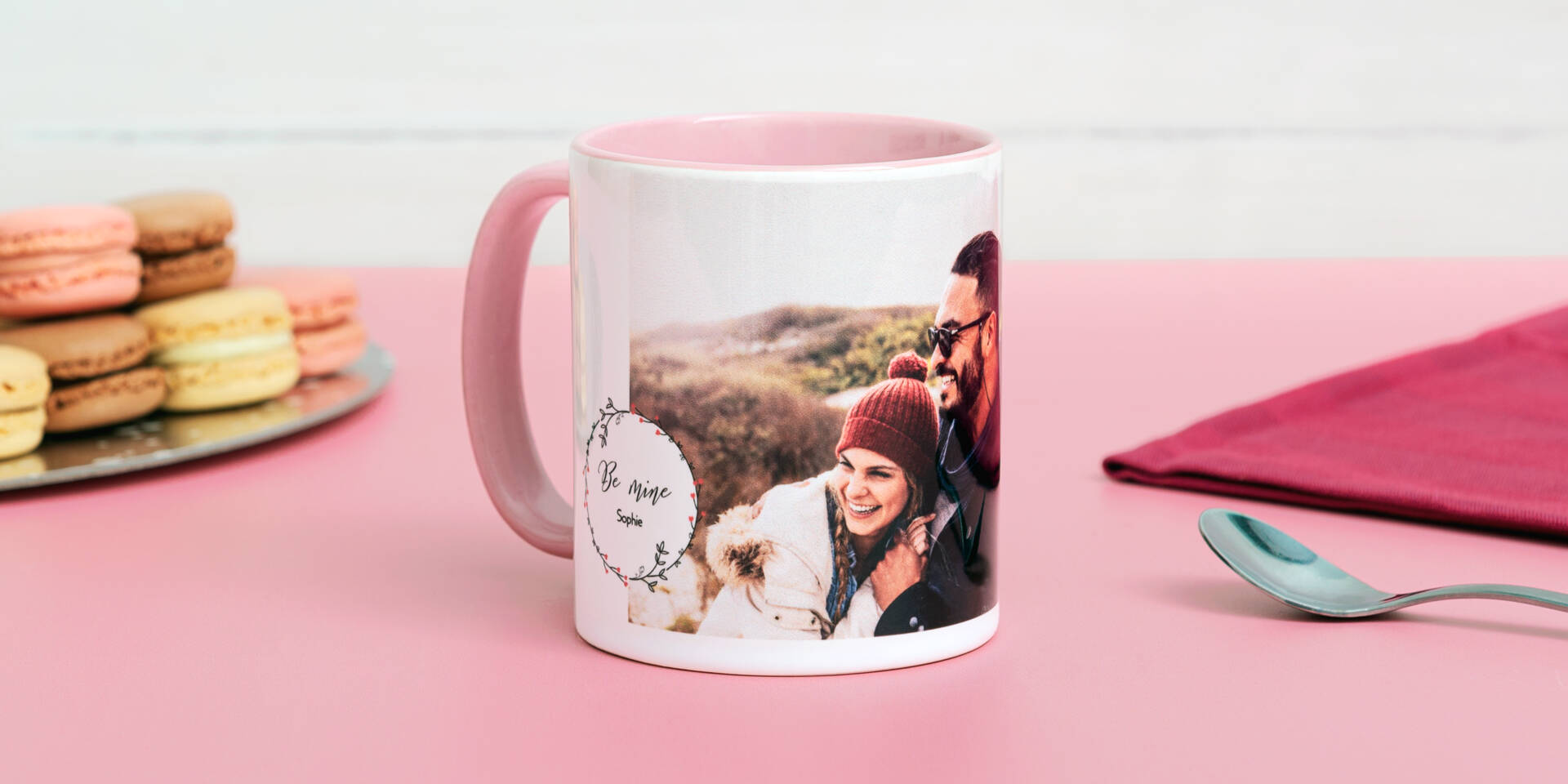 Roze mok met foto van een lachend stel, 'Be Mine Sophie' tekst en macarons op een roze tafel.