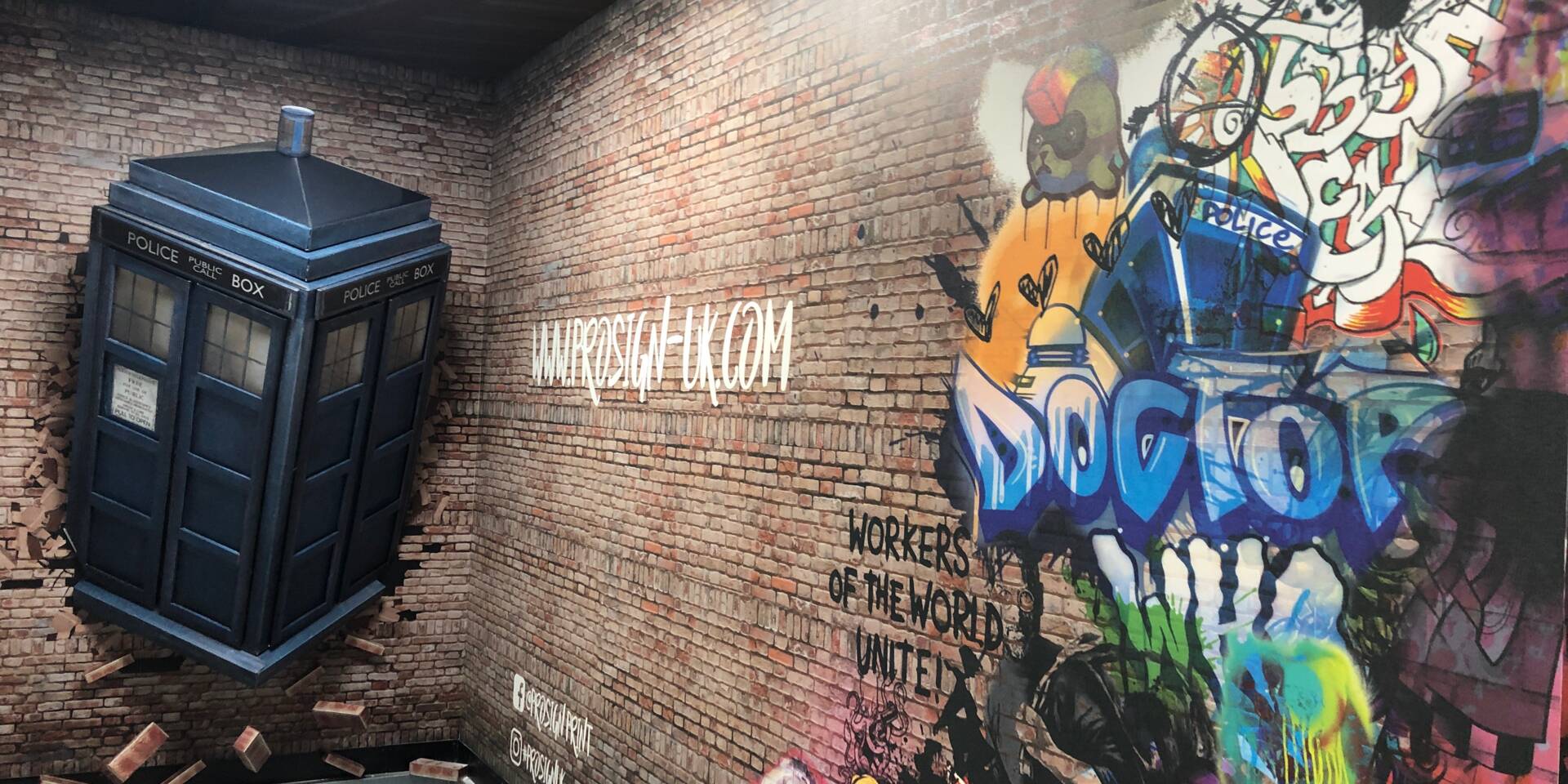 A blue police public call box, resembling the TARDIS, breaks through a brick wall with graffiti art featuring 'DOCTOR' and 'WORKERS OF THE WORLD UNITE'.