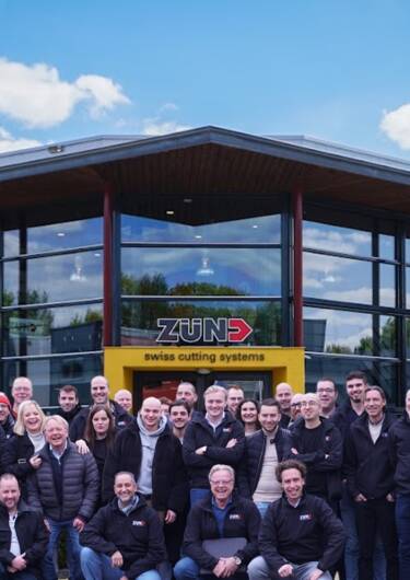 Het Zünd team staat trots opgesteld in een moderne werkplaats, omringd door snijsystemen en bedrijfsinrichting.