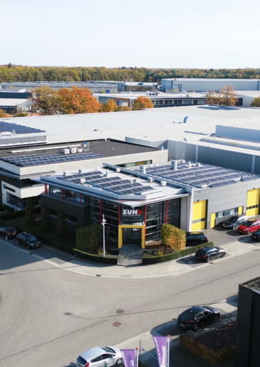 Zünd Systemtechnik gebouw met gele ingang en grote ramen, onder een blauwe lucht met wolken.