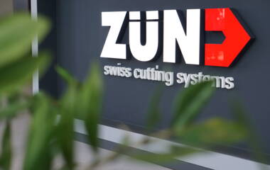 Zünd Systemtechnik logo op een donkergrijze muur in een moderne receptie met rode fauteuils.