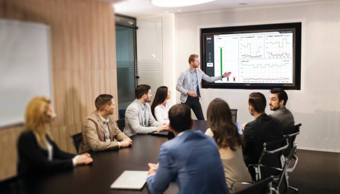 A presenter points to graphs on a screen during a business meeting with colleagues seated around a table. A presenter points to graphs on a screen during a business meeting with colleagues seated around a table.