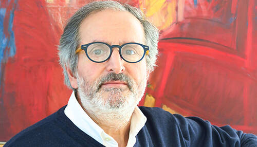 Ritratto di Pino Perri con occhiali e maglione blu, davanti a uno sfondo astratto rosso e giallo.