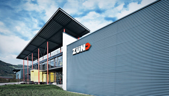 The modern Zünd Systemtechnik headquarters building features corrugated metal siding and a prominent illuminated Zünd logo against a blue sky. The modern Zünd Systemtechnik headquarters building features corrugated metal siding and a prominent illuminated Zünd logo against a blue sky.