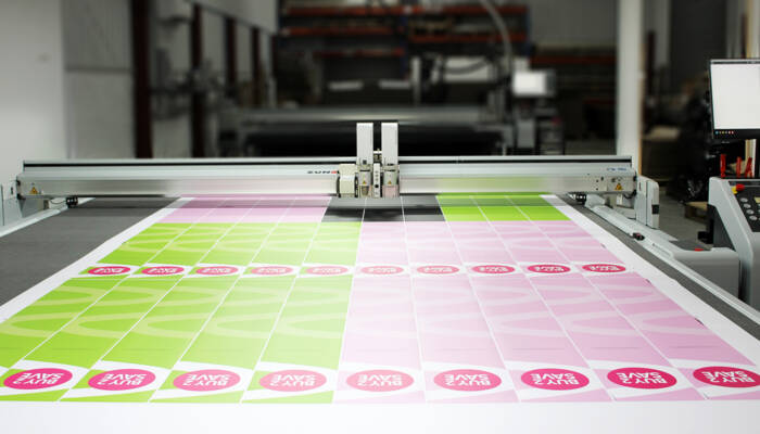 A Zünd UPG3 digital cutting system precisely cuts large sheets with 'BUY 2 SAVE' graphics, showcasing efficient production. A Zünd UPG3 digital cutting system precisely cuts large sheets with 'BUY 2 SAVE' graphics, showcasing efficient production.