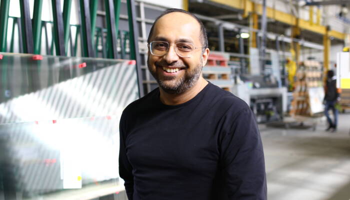 A smiling man with glasses and a beard stands in a Zünd Systemtechnik factory, with large glass panes behind him. A smiling man with glasses and a beard stands in a Zünd Systemtechnik factory, with large glass panes behind him.