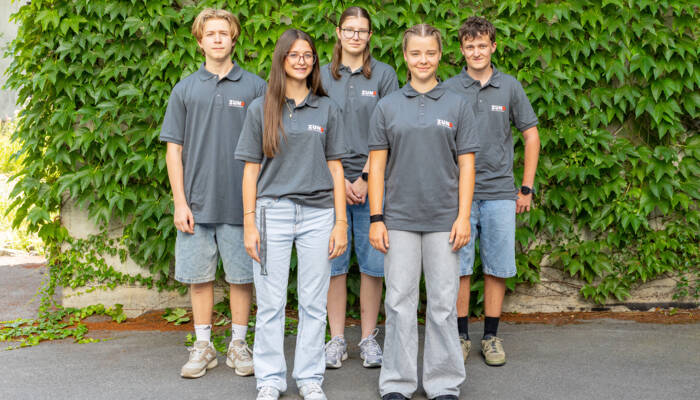 Im Sommer posieren sieben neue Lernende von Z&uuml;nd Systemtechnik in grauen Poloshirts vor einem modernen Geb&auml;ude.
