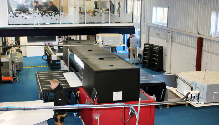 Zünd UPG4 digital cutting machine on a factory floor with workers and other industrial equipment in the background. Zünd UPG4 digital cutting machine on a factory floor with workers and other industrial equipment in the background.