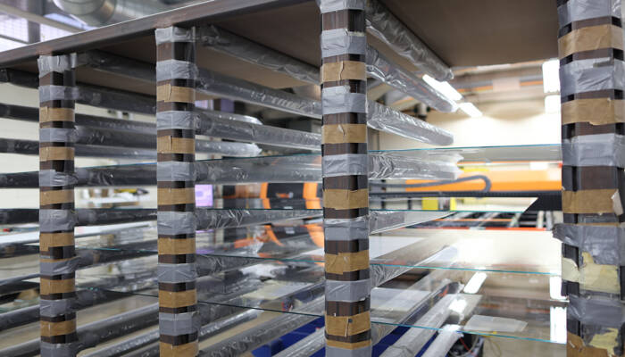 Glass sheets protected by plastic wrap are stored on a metal rack with taped supports in a manufacturing facility. Glass sheets protected by plastic wrap are stored on a metal rack with taped supports in a manufacturing facility.