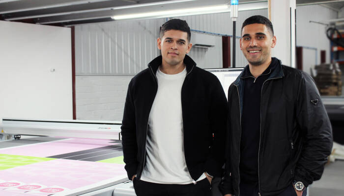 Two men stand in front of a Zünd cutting system with a large format print. Two men stand in front of a Zünd cutting system with a large format print.