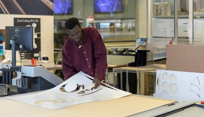 Man in maroon shirt examining a large sheet with intricate cutouts, likely from a Zünd cutting system. Man in maroon shirt examining a large sheet with intricate cutouts, likely from a Zünd cutting system.