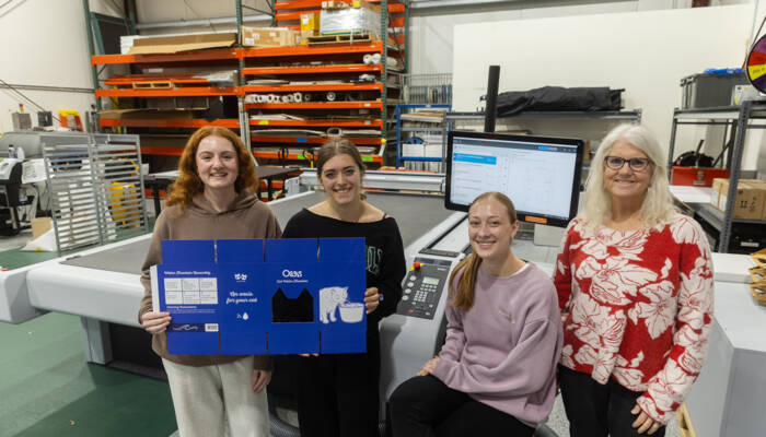 Team presents finished packaging prototype for a cat water fountain beside cutting machine in a production workshop.