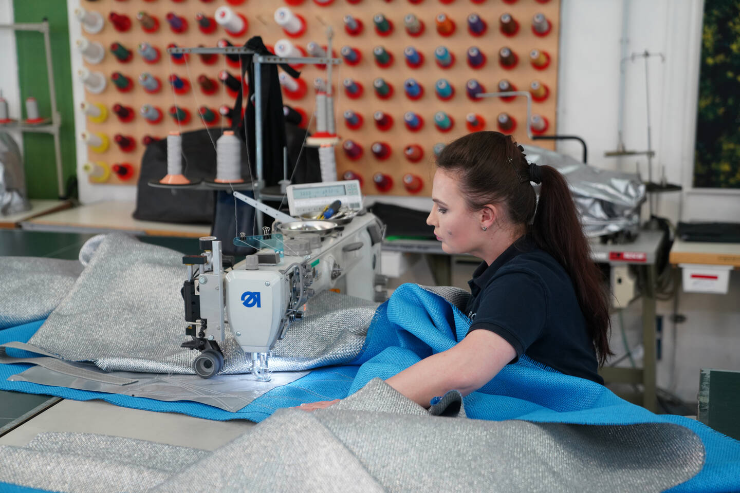 A woman operates a Zünd sewing machine, meticulously stitching together metallic silver and bright blue fabrics in a workshop. A woman operates a Zünd sewing machine, meticulously stitching together metallic silver and bright blue fabrics in a workshop.