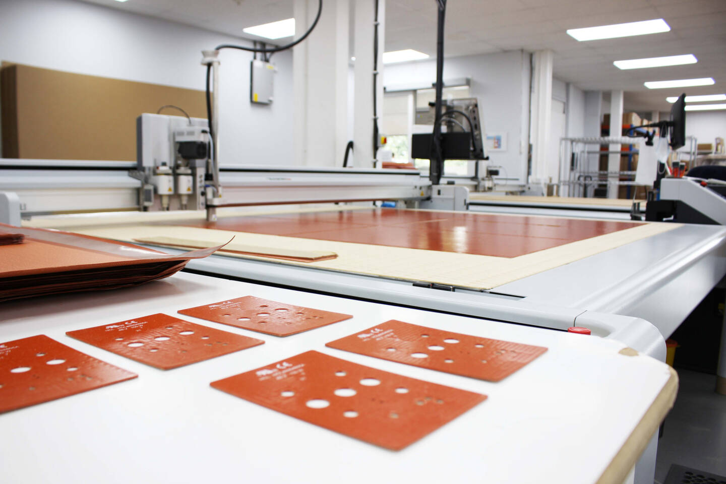 Zünd cutting system processing orange silicone sheets with pre-cut holes, ready for assembly on a workbench. Zünd cutting system processing orange silicone sheets with pre-cut holes, ready for assembly on a workbench.