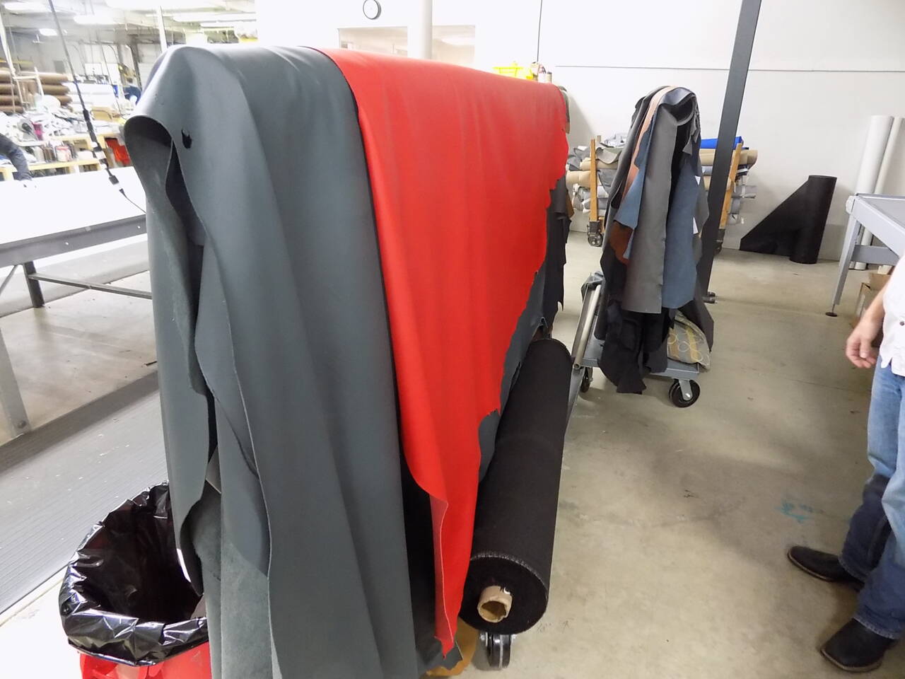 Rolls of dark grey and red leather are displayed next to a cart with fabric samples in a workshop setting. Rolls of dark grey and red leather are displayed next to a cart with fabric samples in a workshop setting.