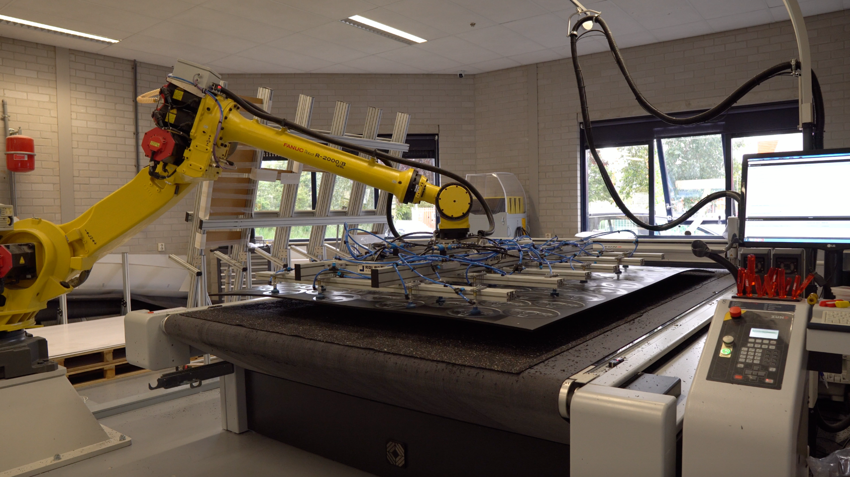 Gele FANUC R-2000iB robotarm boven een Z&uuml;nd snijtafel met zuignappen die een materiaal vasthouden.