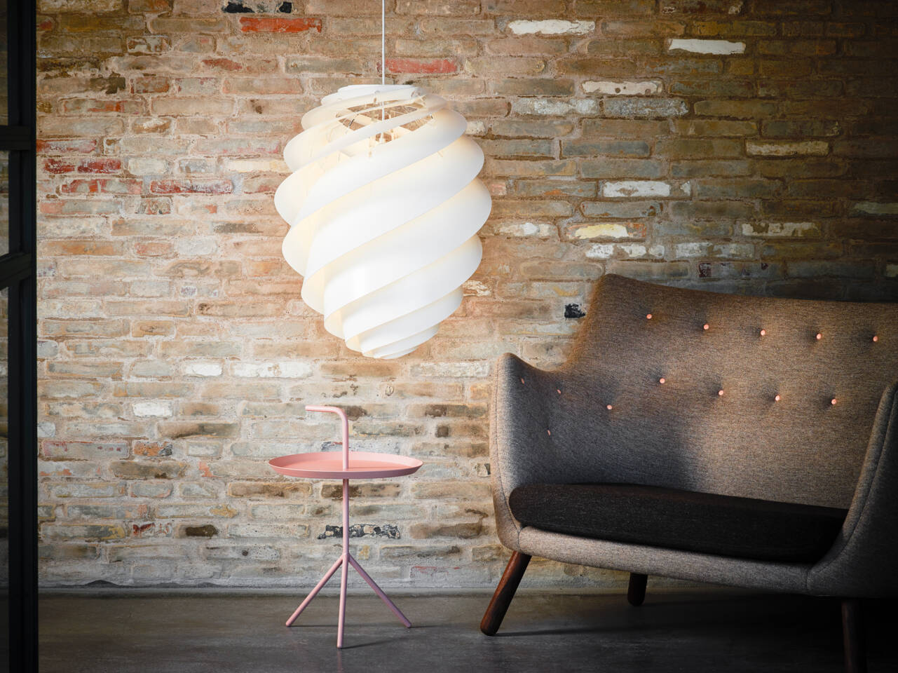 A white spiral pendant light hangs above a small pink side table next to a tufted grey armchair against a brick wall.