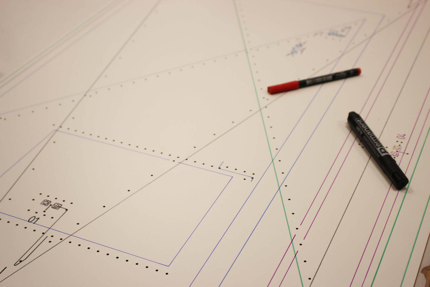 Two permanent markers, one red and one black, rest on a technical drawing with lines and dots, suggesting design or planning. Two permanent markers, one red and one black, rest on a technical drawing with lines and dots, suggesting design or planning.