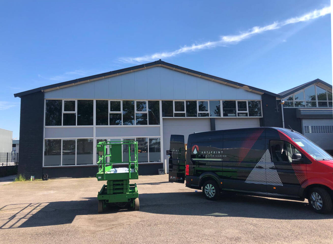 Een groene hoogwerker staat voor een gebouw met grote ramen en een bestelwagen met het opschrift 'ARTIPRINT'.