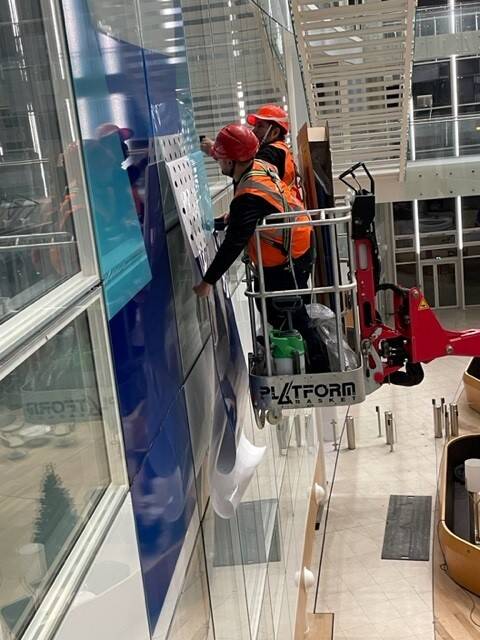 Deux ouvriers en nacelle installent des panneaux bleus sur la façade vitrée d'un bâtiment moderne.