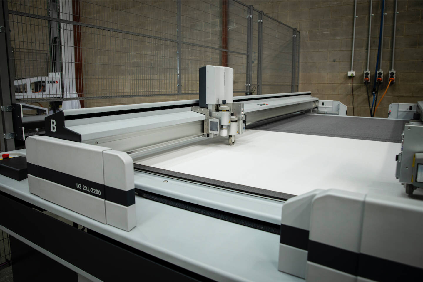 A Zünd D3 2XL-3200 digital cutting system with a cutting head positioned over a large white material on a grey cutting mat. A Zünd D3 2XL-3200 digital cutting system with a cutting head positioned over a large white material on a grey cutting mat.