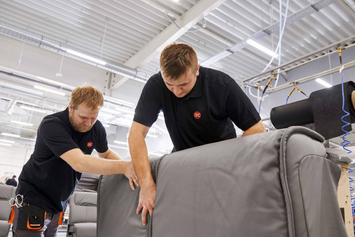 Mitarbeiter der 3C-Gruppe montieren Polsterteile in einer Fabrikhalle mit Zünd Systemtechnik Geräten im Hintergrund. Mitarbeiter der 3C-Gruppe montieren Polsterteile in einer Fabrikhalle mit Zünd Systemtechnik Geräten im Hintergrund.
