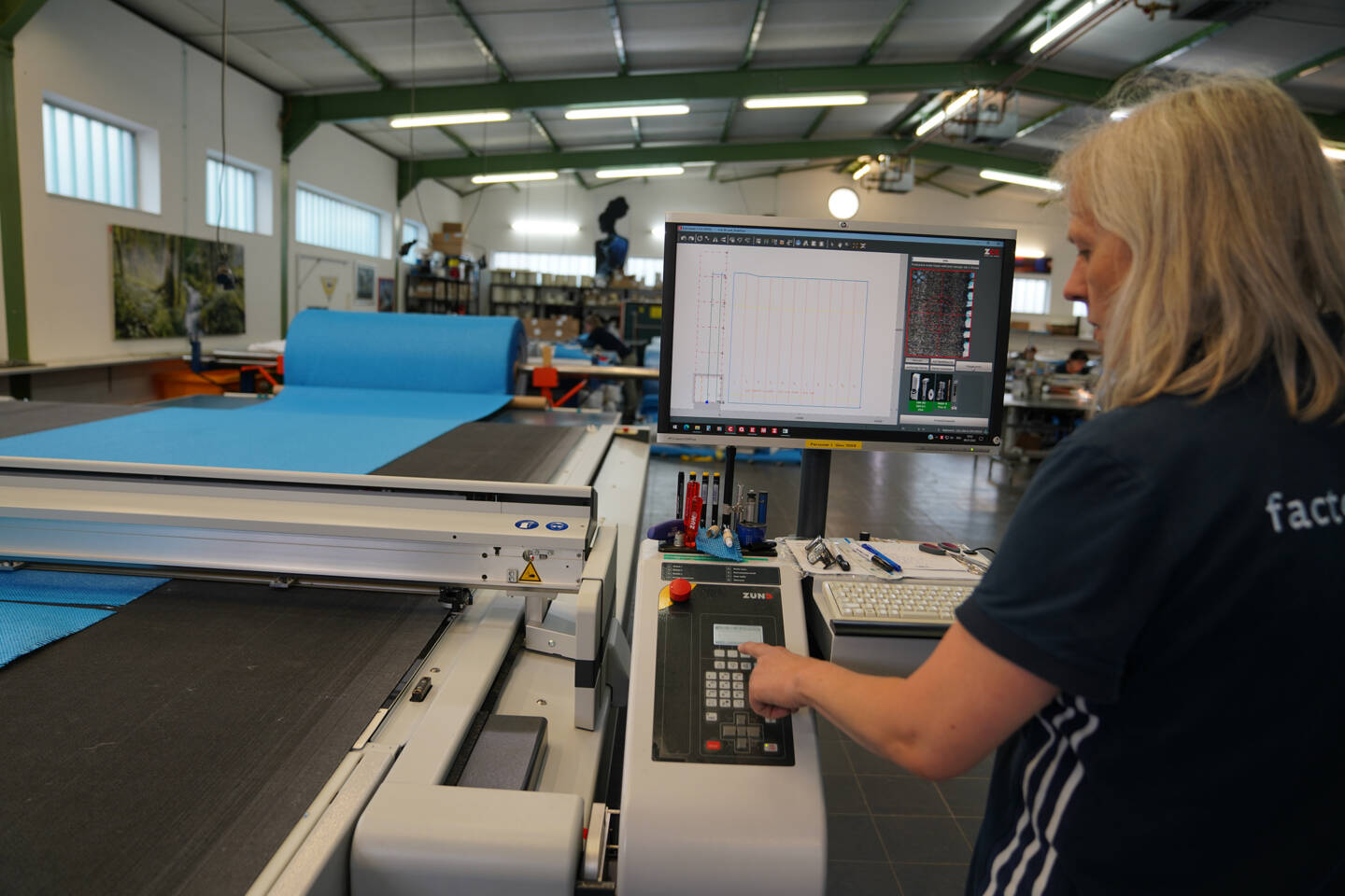 A woman operates a Zünd cutting machine, with a large roll of blue material and a computer interface displaying cutting patterns. A woman operates a Zünd cutting machine, with a large roll of blue material and a computer interface displaying cutting patterns.