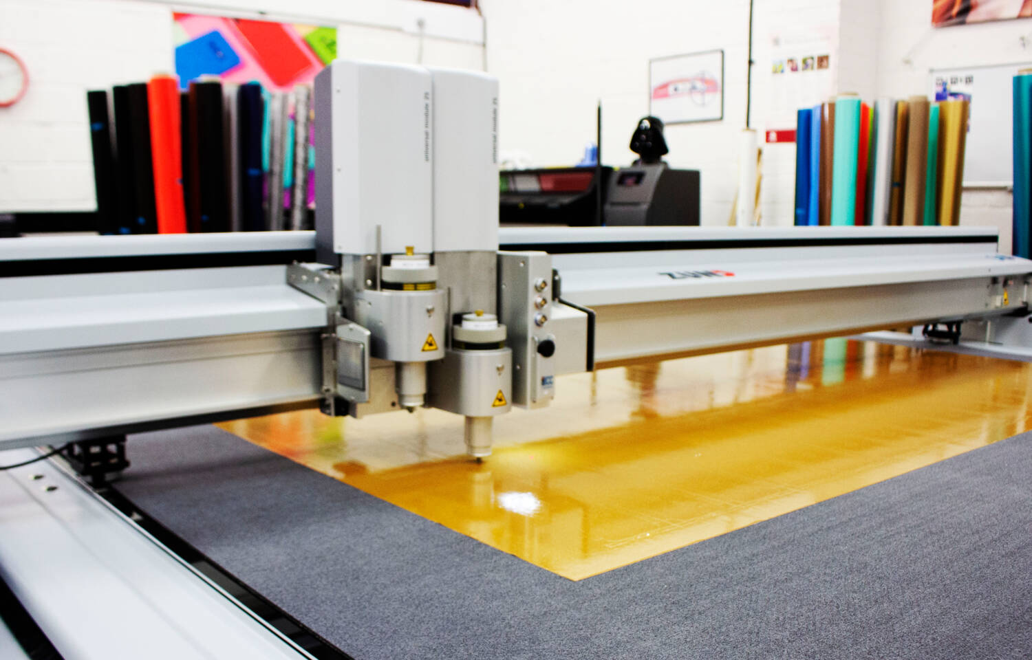 Zünd G3 L-2500 digital cutter precisely scores a large sheet of reflective orange material on a work surface. Zünd G3 L-2500 digital cutter precisely scores a large sheet of reflective orange material on a work surface.