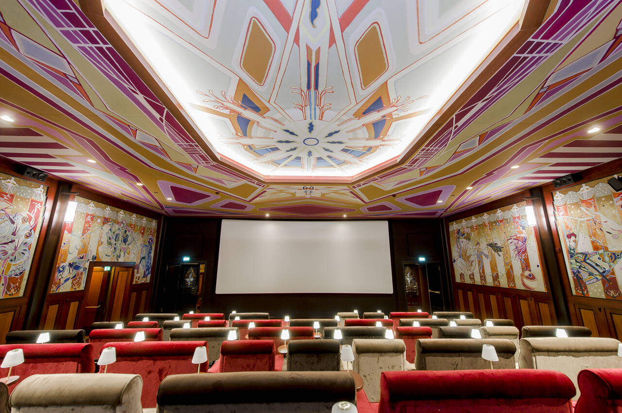 Gedetailleerd interieur van een bioscoop met art-deco plafond en muurschilderingen, witte projectiescherm en rode stoelen.