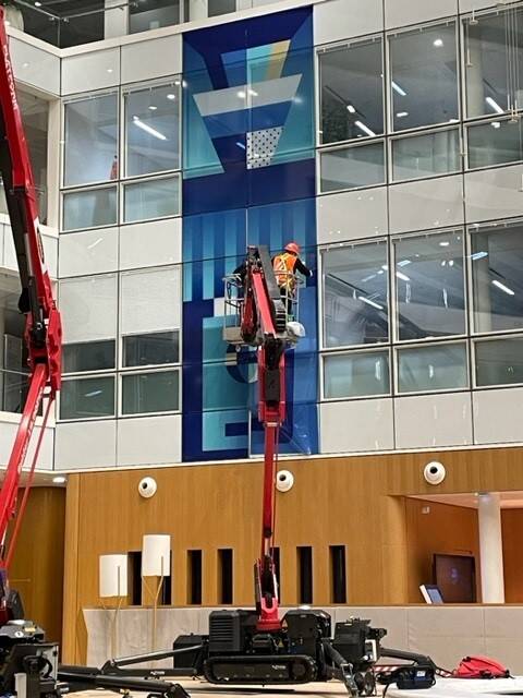 Travailleurs dans une nacelle élévatrice installent une grande œuvre d'art graphique sur la façade vitrée d'un bâtiment.
