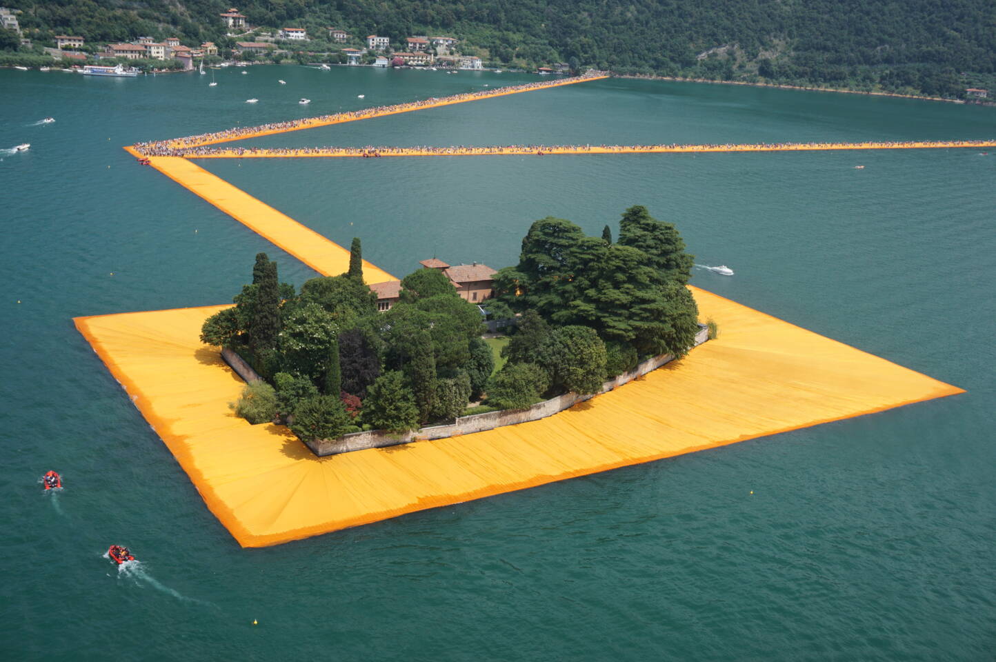 The Floating Piers installation on Lake Iseo, Italy, featuring vibrant orange walkways connecting islands and the shore, with people walking. The Floating Piers installation on Lake Iseo, Italy, featuring vibrant orange walkways connecting islands and the shore, with people walking.