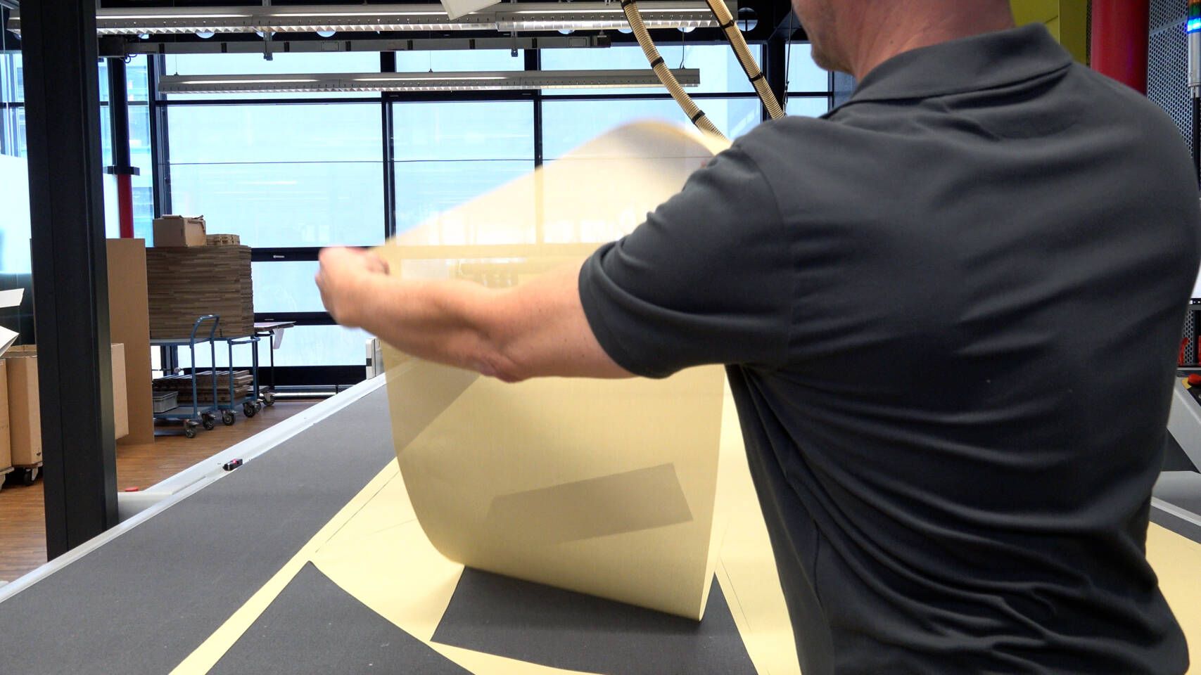 A worker holds a large sheet of translucent material over a cutting table, preparing it for processing in a Zünd cutting system. A worker holds a large sheet of translucent material over a cutting table, preparing it for processing in a Zünd cutting system.