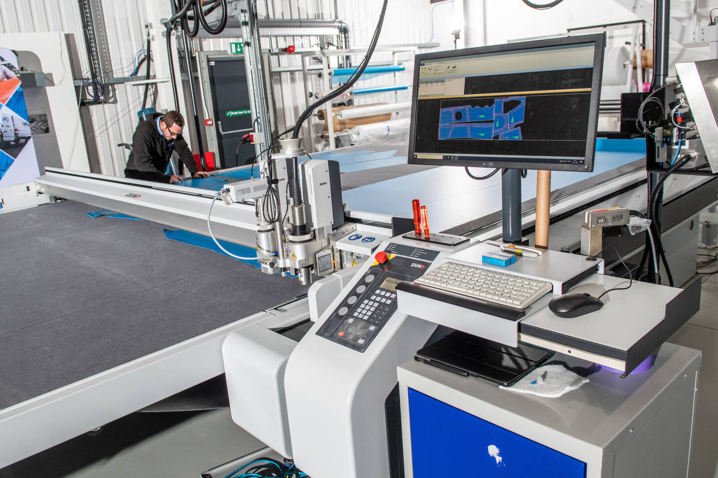 A man operates a Zünd G3 digital cutting system, with a computer displaying cutting patterns and a keyboard nearby. A man operates a Zünd G3 digital cutting system, with a computer displaying cutting patterns and a keyboard nearby.