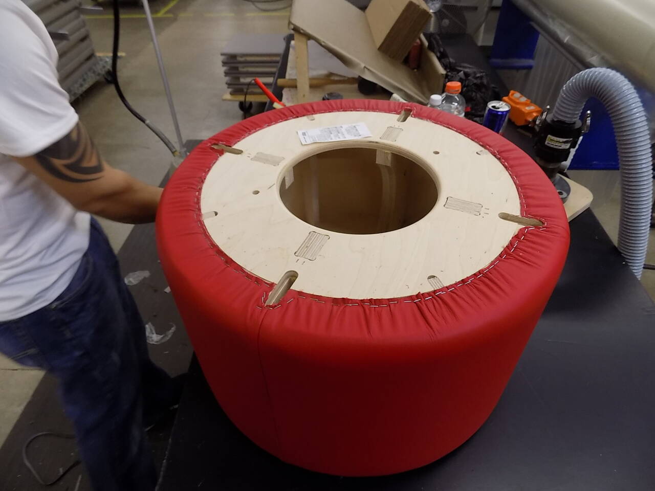A person in a white shirt and blue jeans is working on a red upholstered cylindrical object with a wooden top. A person in a white shirt and blue jeans is working on a red upholstered cylindrical object with a wooden top.
