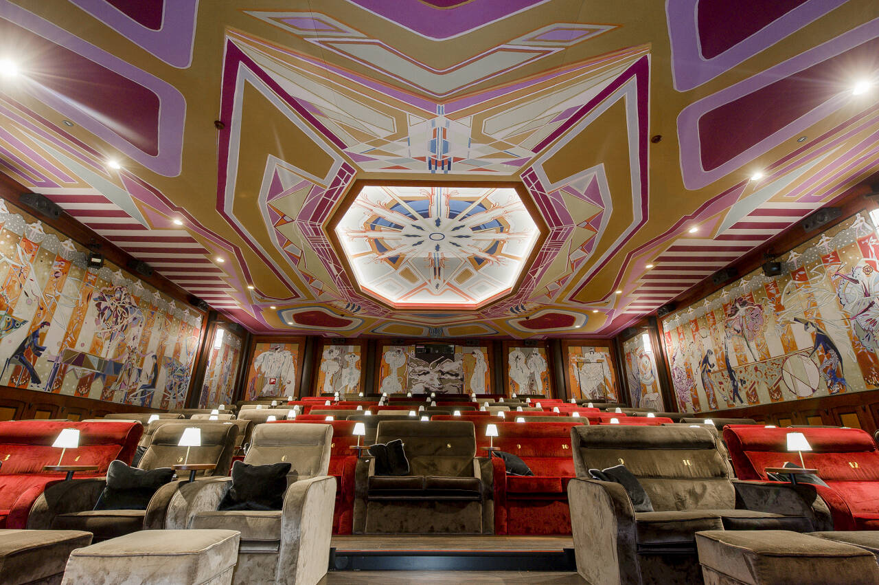 Luxe bioscoopzaal met art-deco plafond, muurschilderingen en comfortabele stoelen in rood en bruin.
