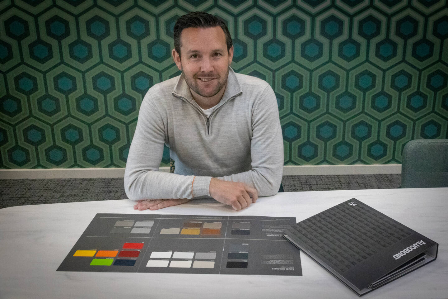 Man in grijs gebreid vest zit achter een tafel met een Alucobond kleurenpalet en monstertafel.