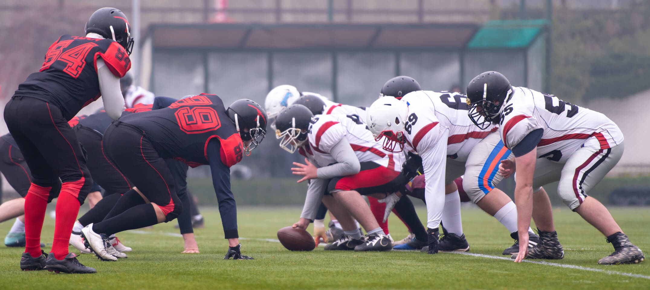 Zwei American-Football-Teams in voller Montur in Startposition auf einem Rasenfeld, bereit zum Ansto&szlig;.