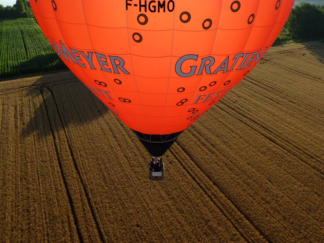 Orange hot air balloon with 'MEY ER GRATIE' text flying over a harvested wheat field, with people in the basket. Orange hot air balloon with 'MEY ER GRATIE' text flying over a harvested wheat field, with people in the basket.