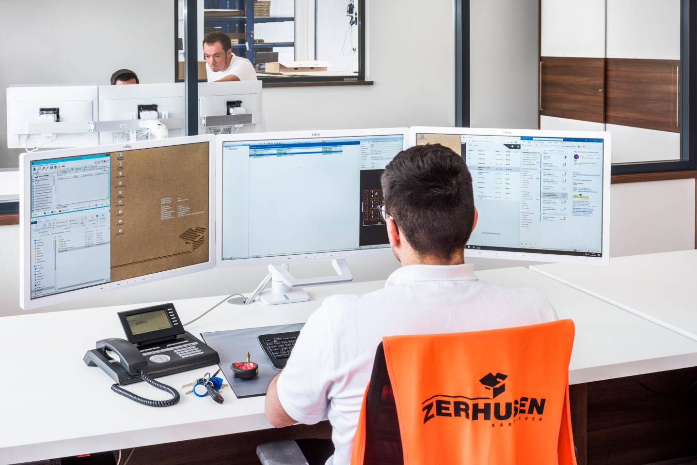 Mitarbeiter arbeitet an einem Schreibtisch mit drei Monitoren und einem Telefon.