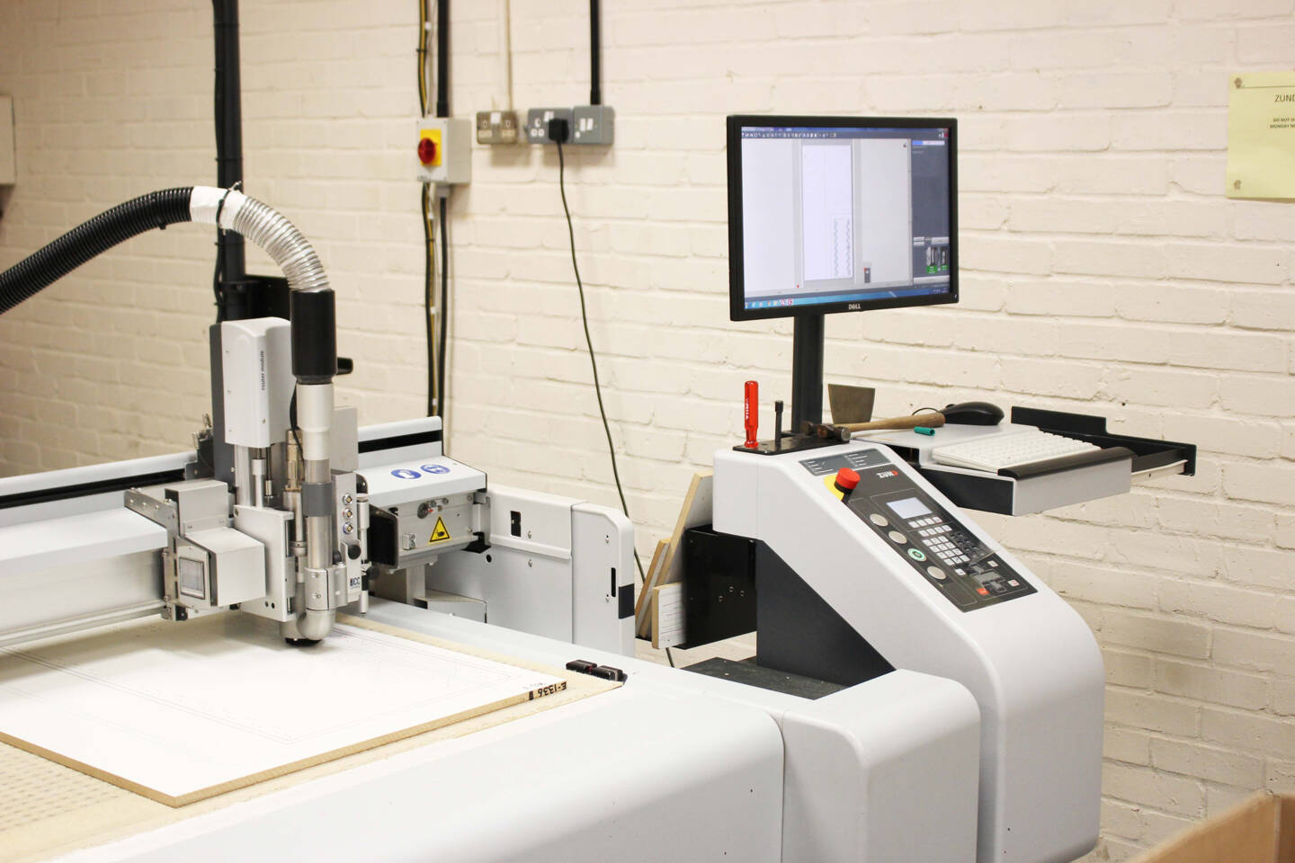 Zünd cutting system with router module, computer monitor displaying design software, keyboard, and tools in a workshop setting. Zünd cutting system with router module, computer monitor displaying design software, keyboard, and tools in a workshop setting.