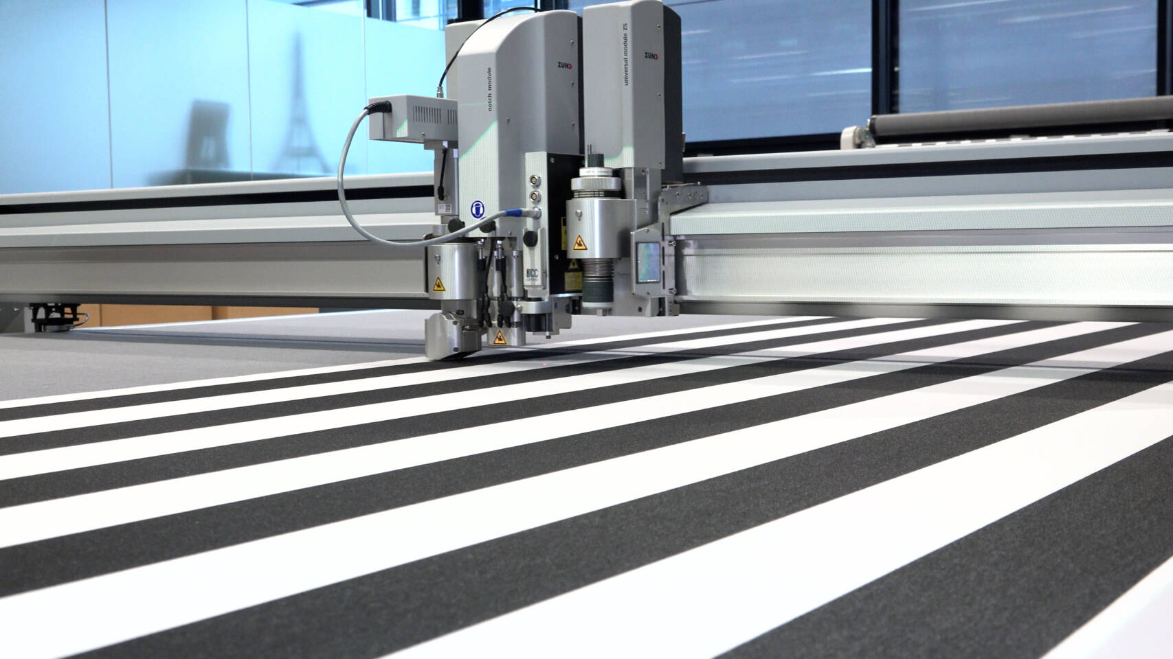Zünd cutting system with a universal module precisely cutting black and white striped material on a flatbed. Zünd cutting system with a universal module precisely cutting black and white striped material on a flatbed.