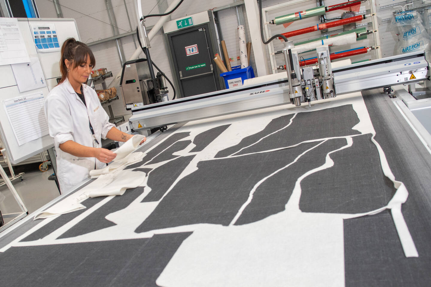 A woman in a lab coat inspects fabric pieces laid out on a Zünd cutting machine bed in a manufacturing facility. A woman in a lab coat inspects fabric pieces laid out on a Zünd cutting machine bed in a manufacturing facility.