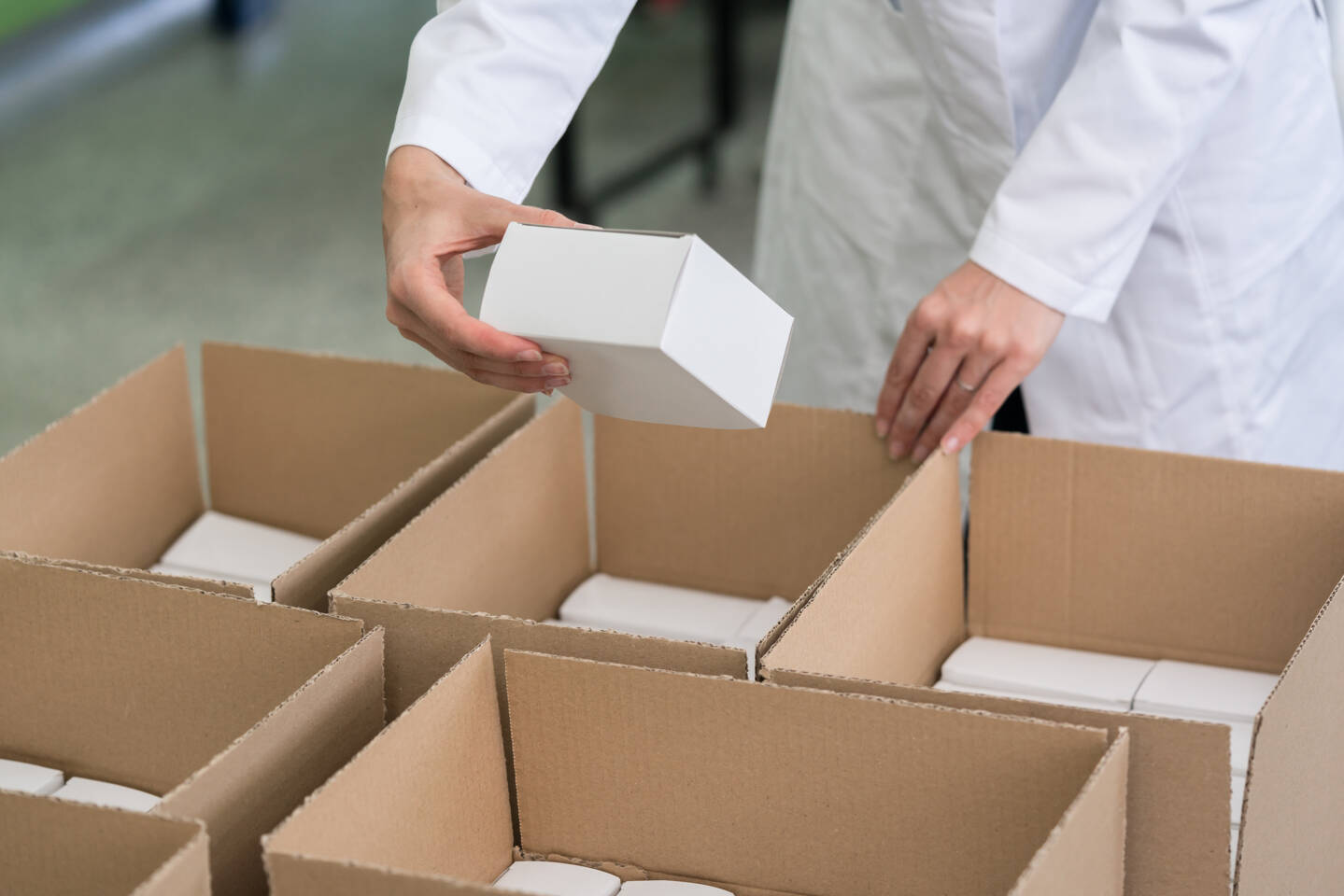 Une main en blouse blanche place une petite bo&icirc;te blanche dans une bo&icirc;te en carton lors de la production en petite s&eacute;rie.