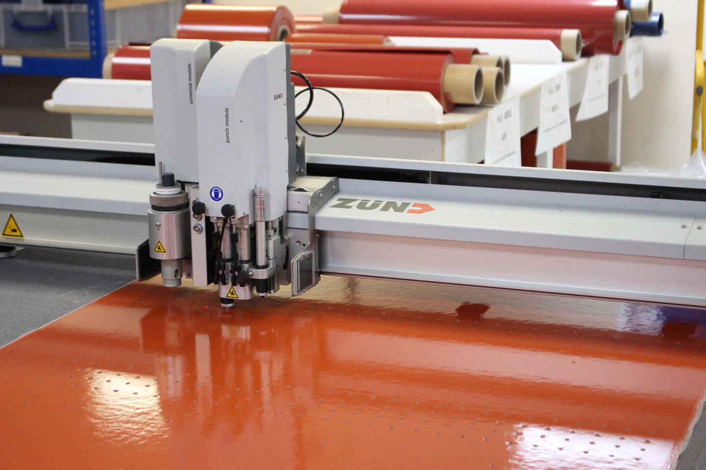 A Z&uuml;nd G3 digital cutting system with a punch module precisely perforates an orange, high-gloss material.